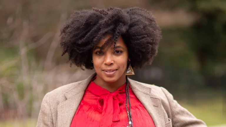 A confident woman with voluminous natural curly hair, wearing a red blouse with a bow detail and a beige blazer, standing outdoors with a soft-focus natural background.