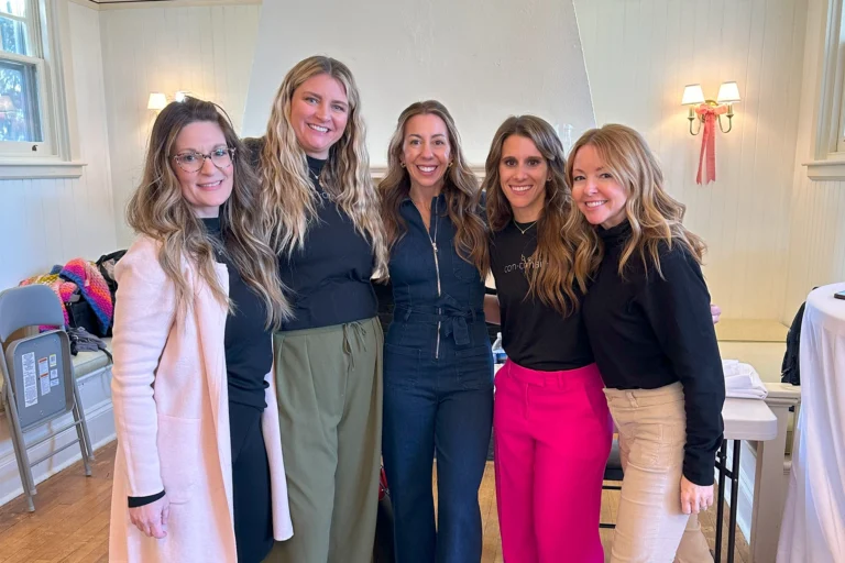 A group of five smiling women posing together in a warmly lit room. Two of them are wearing “Concihairge” branded black tops. They stand close together, dressed in stylish outfits, with a cozy and welcoming atmosphere in the background.